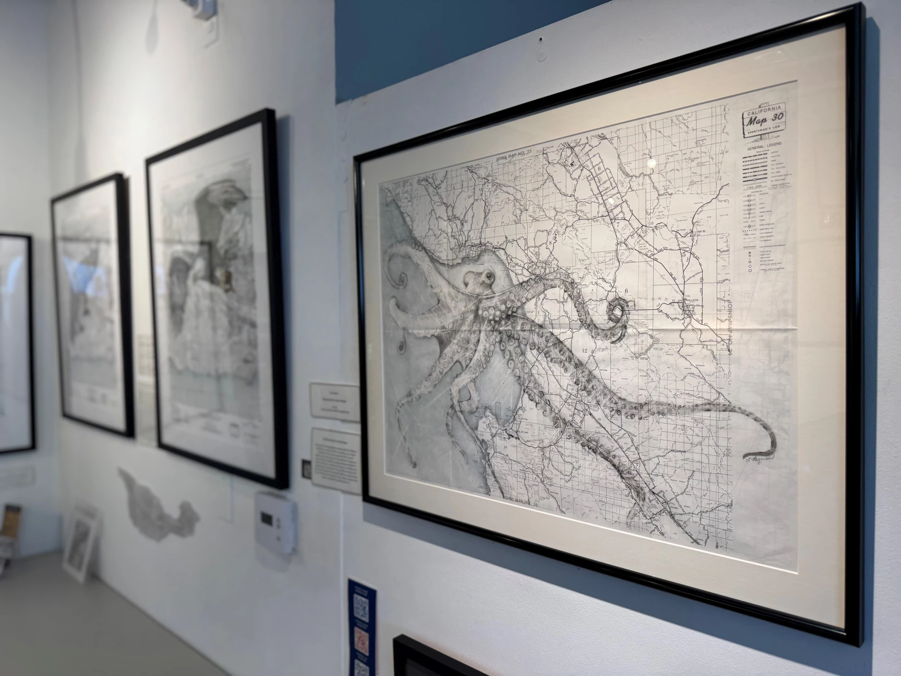 Several pieces of art hang on a gallery wall. 
          One piece, in focus, depicts an octopus overlaid on a map.